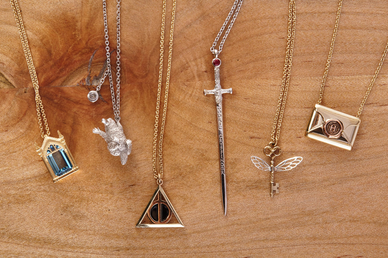 Selection of six necklaces in Officially licensed Harry Potter fine jewelry collection by Freeman Design on a rustic wooden background. Featured items: Mirror of Erised in 14k yellow gold, Niffler in sterling silver, Deathly Hallows in 14k yellow gold, Sword of Gryffindor in platinum, Winged Key in 18k yellow gold, and Hogwarts Acceptance Letter in 14k yellow gold. 