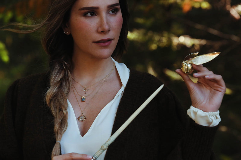 Model wearing 14k gold winged key necklace, mirror of erised necklace, time turner necklace and holding a golden snitch ring box