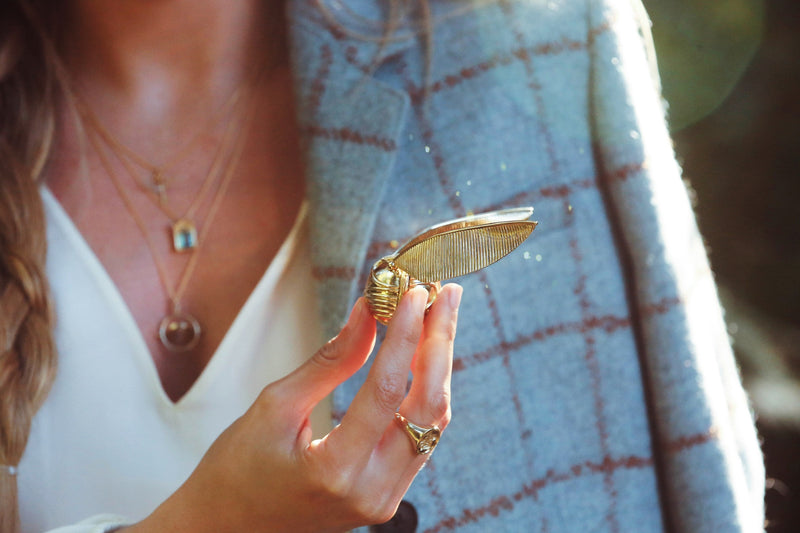 Close view of a model’s hand holding an officially licensed Harry Potter Golden Snitch Ring Box and wearing a Hogwarts Crest Signet Ring in solid 14k yellow gold. Also available in other metals. 